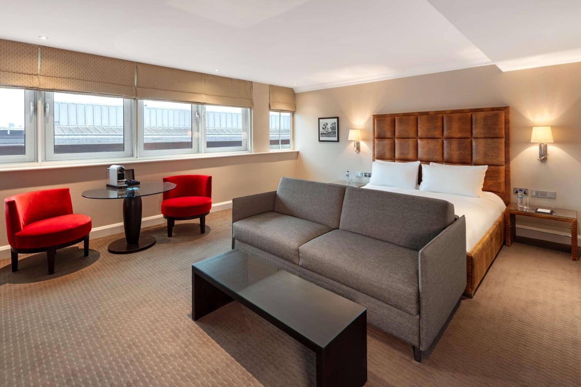 The May Fair, A Radisson Collection Hotel, Mayfair لندن المظهر الخارجي الصورة The photo shows a modern hotel room featuring a comfortable setup. There is a large double bed with a headboard, some decorative artwork on the wall, and two windows allowing natural light to enter. In the room, there's a gray sofa facing a dark coff