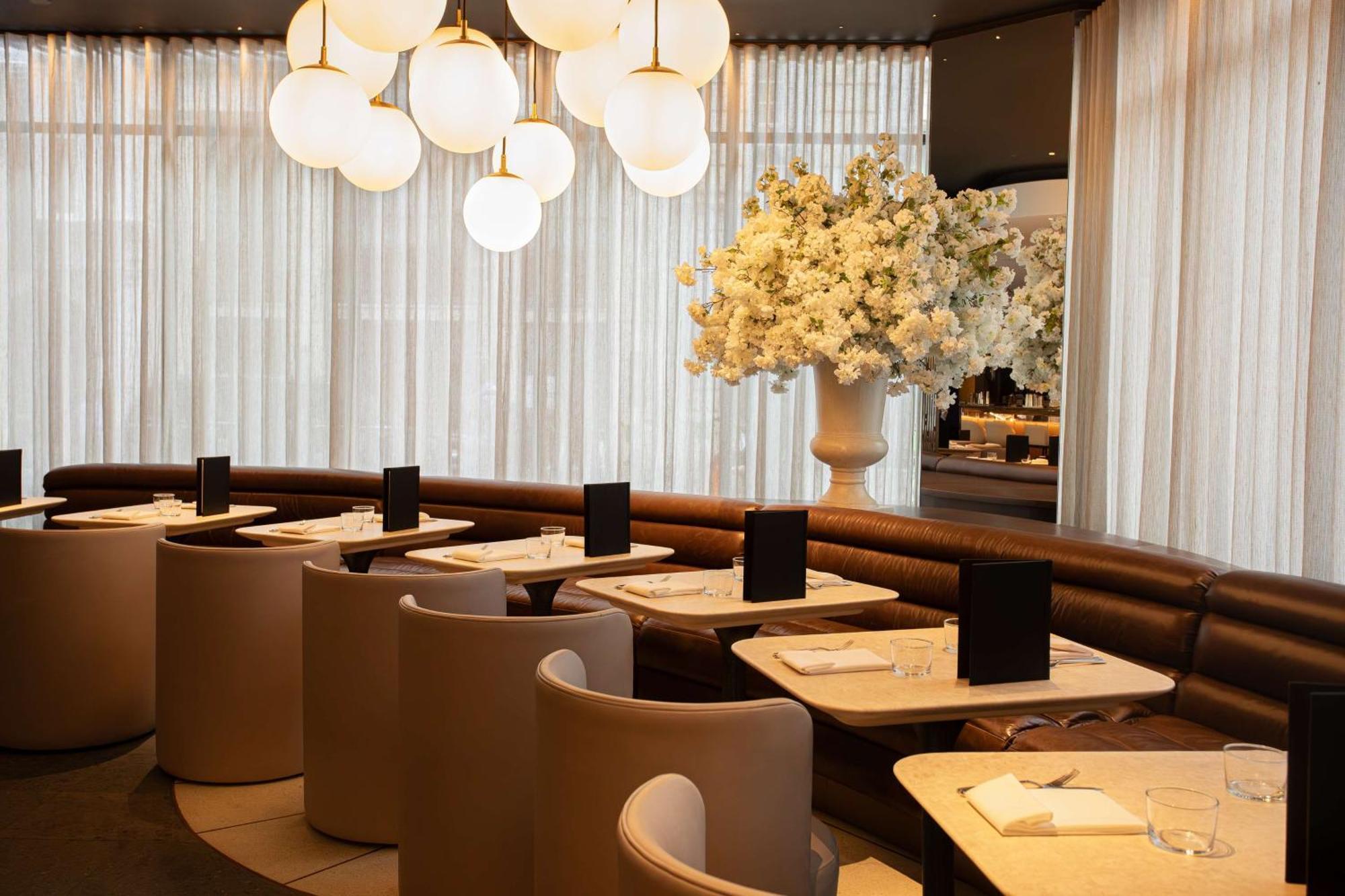 The May Fair, A Radisson Collection Hotel, Mayfair لندن المظهر الخارجي الصورة The photo shows a modern restaurant interior. It features stylish seating arrangements with curved booths and elegant tables. The lighting is soft, provided by large, round pendant lights hanging from the ceiling. A large floral arrangement in a whit